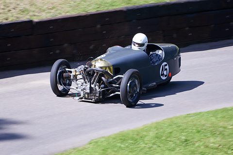 A dark green racing three-wheeler with an exposed V-twin at its nose and a helmeted driver, on a tarmac bend by a grass bank. Opens a larger view