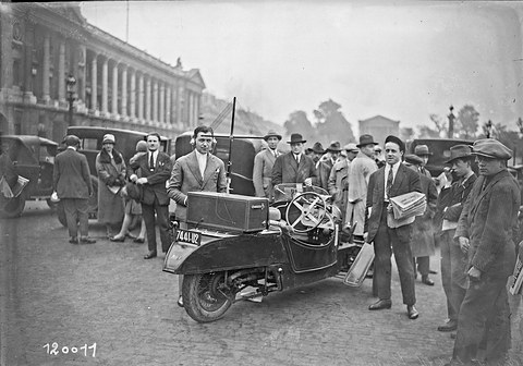 A small open three-wheeler with a box-shaped radio set on its tail and a mast aerial, a man in headphones and a crowd round it. Opens a larger view