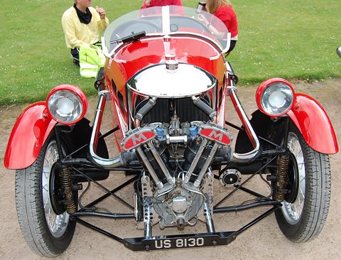 Front view of a red three-wheeler with a chrome cowl, an exposed V-twin with M badges and two headlamps. Opens a larger view