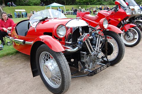 A red three-wheeler with exposed V-twin and chrome cowl beside red modern motorcycles on gravel. Opens a larger view