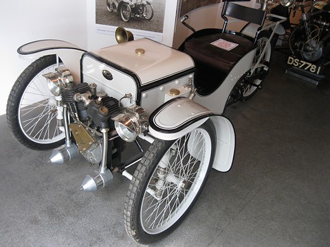 A white early three-wheeler with an exposed V-twin, brass horn and leather seat on a grey floor. Opens a larger view