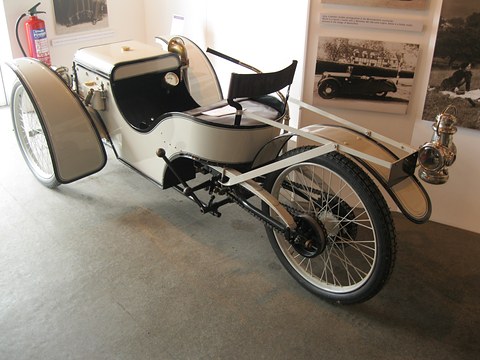 Side view of a white early three-wheeler with a box bonnet and open tubular frame, framed photographs behind. Opens a larger view