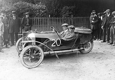 Two men in caps in a dark three-wheeler numbered 20 with exposed engine, a crowd of men behind a fence.