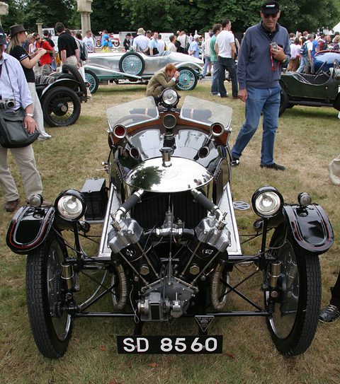 Front view of a black three-wheeler with a polished cowl, exposed V-twin and two headlamps, a crowd on grass behind. Opens a larger view