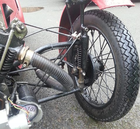 Close view of a front wheel on a vertical pillar with coil springs, tubular frame and exhaust pipe beside it.