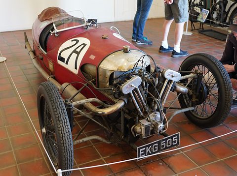 A battered red racing three-wheeler numbered 2A with an exposed V-twin on a tiled floor behind a rope. Opens a larger view
