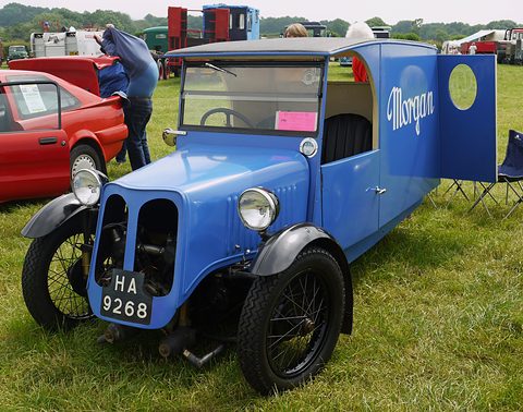 A blue three-wheeled van with a cowled front, two headlamps and Morgan lettered on its open rear door, on grass. Opens a larger view