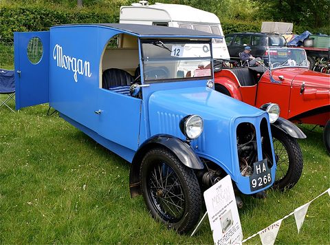 A blue three-wheeled van lettered Morgan with its side door swung open, a placard and bunting at its front wheel. Opens a larger view
