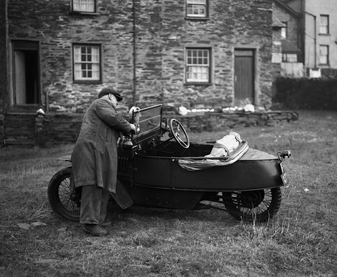 A man in a long coat and cap bends over a small black three-wheeler with wire wheels, parked on grass before slate-stone cottages.