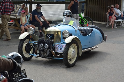 A pale blue three-wheeler with cream wings and a rally plate numbered 196, spectators on benches behind. Opens a larger view