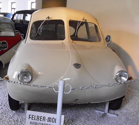 Cream rounded coupé with two headlamps behind a chain barrier, a FELBER-MÖVE sign below. Opens a larger view