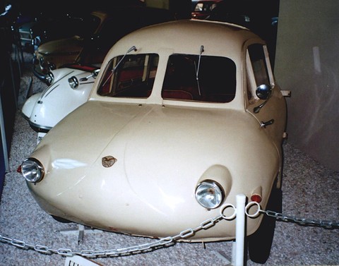 Cream rounded coupé in a dim museum, a white car behind and a chain barrier in front. Opens a larger view