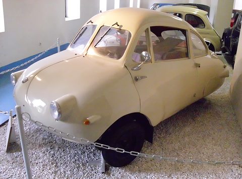 Cream egg-shaped two-door coupé on white gravel inside a museum, chain barrier in front.