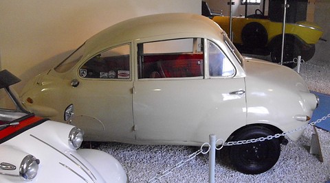Cream rounded coupé side-on behind a chain barrier, red interior visible through the door glass. Opens a larger view