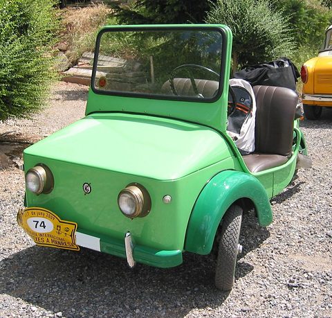 Green open three-wheeler with two round headlamps, a windscreen and a rally plate numbered 74, parked on gravel.