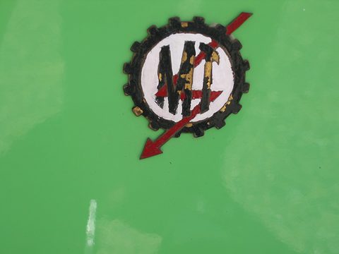 Round badge with MT letters inside a black cog, crossed by a red arrow, on bright green paint. Opens a larger view