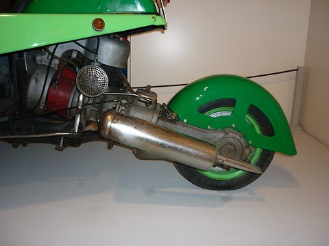 Close view of an exposed engine with a perforated air filter and long silencer next to a green-mudguarded rear wheel. Opens a larger view