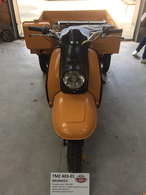 Front of an orange three-wheeled scooter with a single headlamp and a museum label on the floor. Opens a larger view