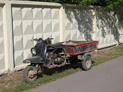 Black three-wheeled cargo scooter with a red box parked on grass before a patterned concrete fence. Opens a larger view