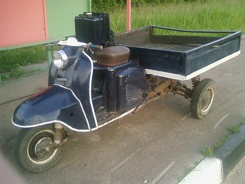 Dark blue three-wheeled scooter with white piping and a flat cargo platform on tarmac.