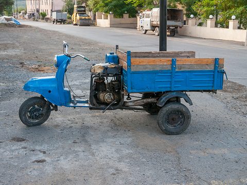 Blue three-wheeled cargo scooter with a wooden load box parked on a gravel verge by a road. Opens a larger view