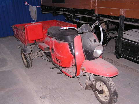 Red three-wheeled cargo scooter with a red load box on paving beside an old railway wagon. Opens a larger view