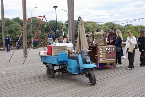 Blue three-wheeled cargo scooter loaded with crates beside a market stall on a wooden boardwalk. Opens a larger view