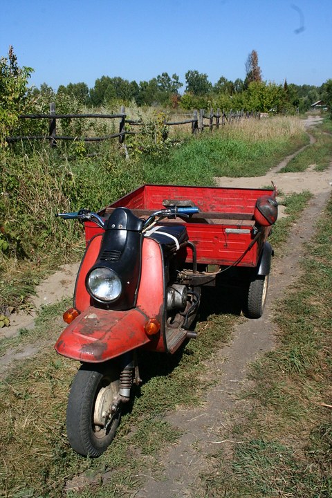 Front of a red three-wheeled scooter with a black leg shield and a red load box on a grassy track. Opens a larger view