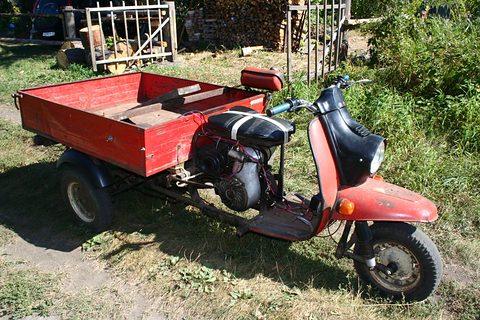 Rear three-quarter of a red three-wheeled scooter with an open engine bay and a red load box. Opens a larger view