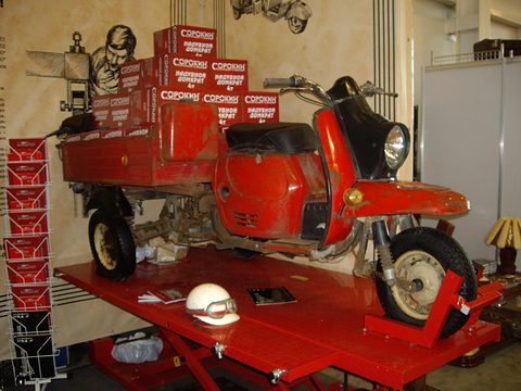 Red three-wheeled cargo scooter with boxes in its load bed on a red lift at a show. Opens a larger view