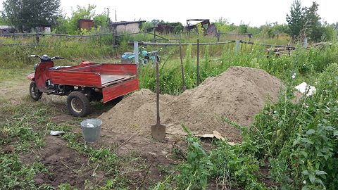 A small red three-wheeled cargo scooter parked by a sand heap and a spade in a vegetable garden.