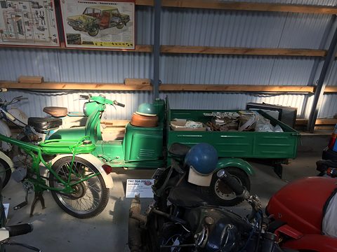 Green three-wheeled cargo scooter among motorcycles and posters in a corrugated-iron museum shed.