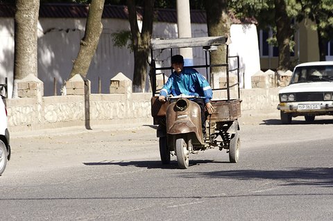 A man in a blue shirt rides a rusty three-wheeled cargo scooter with a cab frame along a road.