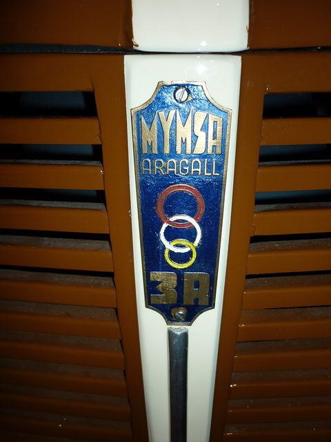 Blue enamel badge reading MYMSA ARAGALL with three coloured rings and 3R, set in a louvred brown grille. Opens a larger view