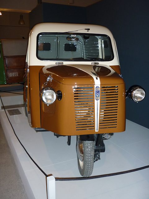 Front of a brown and cream van with a tall louvred grille, two headlamps and a single front wheel, in a dark gallery. Opens a larger view