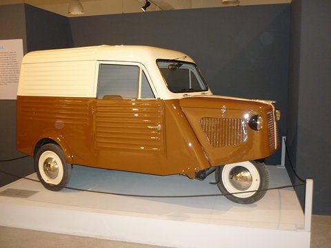Brown and cream three-wheeled van with a louvred nose and ribbed side panels, on a white museum plinth.