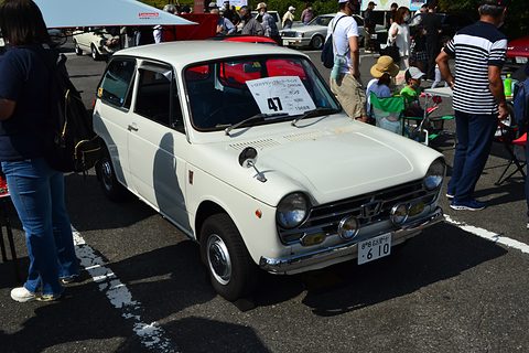 White small saloon with a number card in its windscreen, visitors walking around it. Opens a larger view