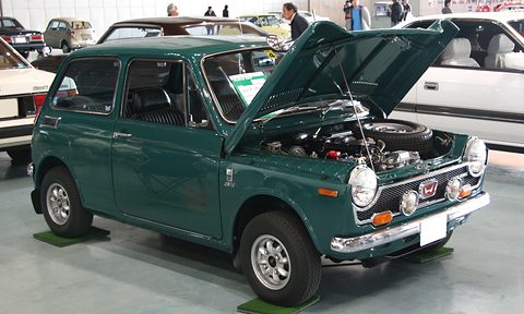 Dark green small saloon with the bonnet up over its engine, parked on mats in a hall. Opens a larger view