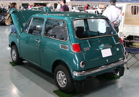 Rear three-quarter of a dark green small saloon with its bonnet raised, visitors behind. Opens a larger view