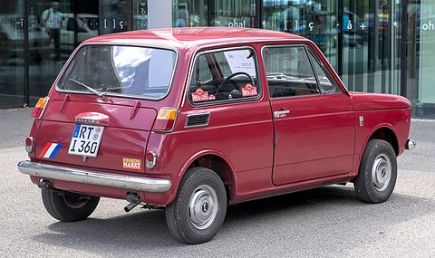 Rear three-quarter of a dark red small saloon with a German plate, a glass building behind. Opens a larger view
