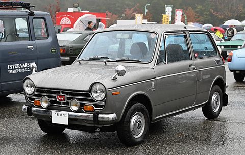 Grey small saloon with fog lamps on wet tarmac, a dark van beside it. Opens a larger view