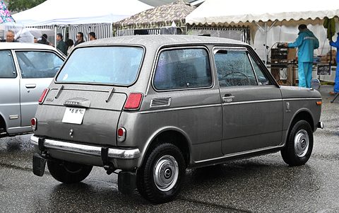 Rear three-quarter of a grey small saloon on wet tarmac, marquees and visitors behind. Opens a larger view