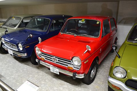 Red small saloon in a museum row between a navy car and a lime-green car. Opens a larger view
