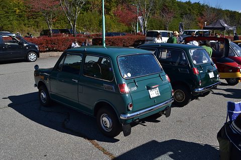 Rear three-quarter of a dark green small saloon among other cars, autumn hills behind. Opens a larger view