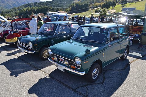 Two dark green small saloons parked on tarmac with visitors and hills behind. Opens a larger view