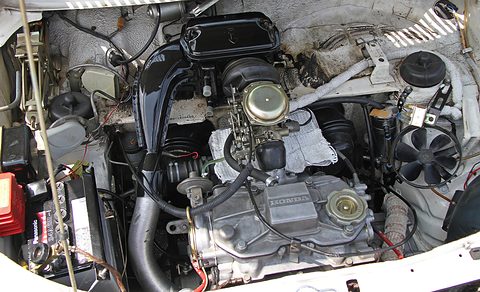 Engine bay crowded with a black air cleaner, a grey cylinder block, wires and a red battery.