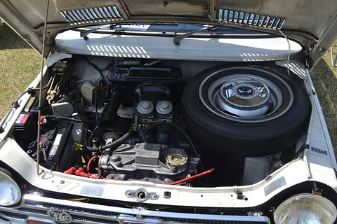 Open bonnet showing a small twin engine with carburettors and a spare wheel beside it. Opens a larger view