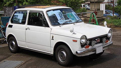 White small saloon with a chequered grille trim and papers in its windscreen, parked under trees. Opens a larger view