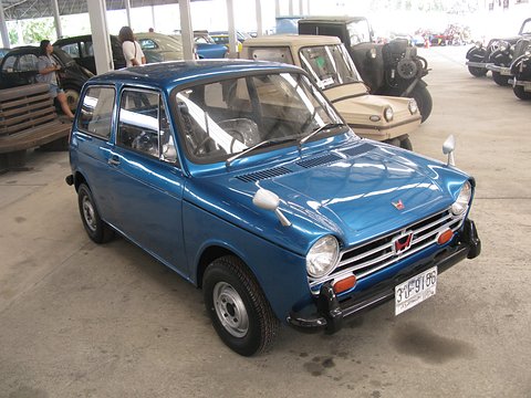 Bright blue small saloon on a concrete floor under an open-sided shed with other old cars. Opens a larger view