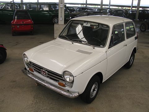 White small saloon on a concrete floor with rows of dark old cars behind. Opens a larger view
